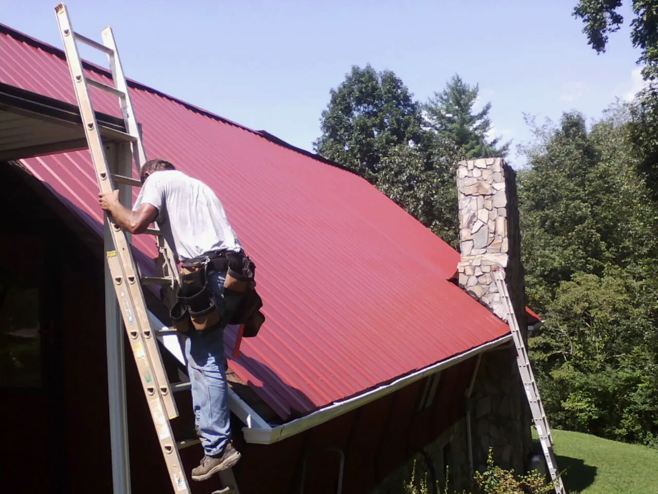 Roofer on a ladder beside a red metal roof and stone chimney — Soffit & Fascia Repair by Roofing Brothers in Spring Church